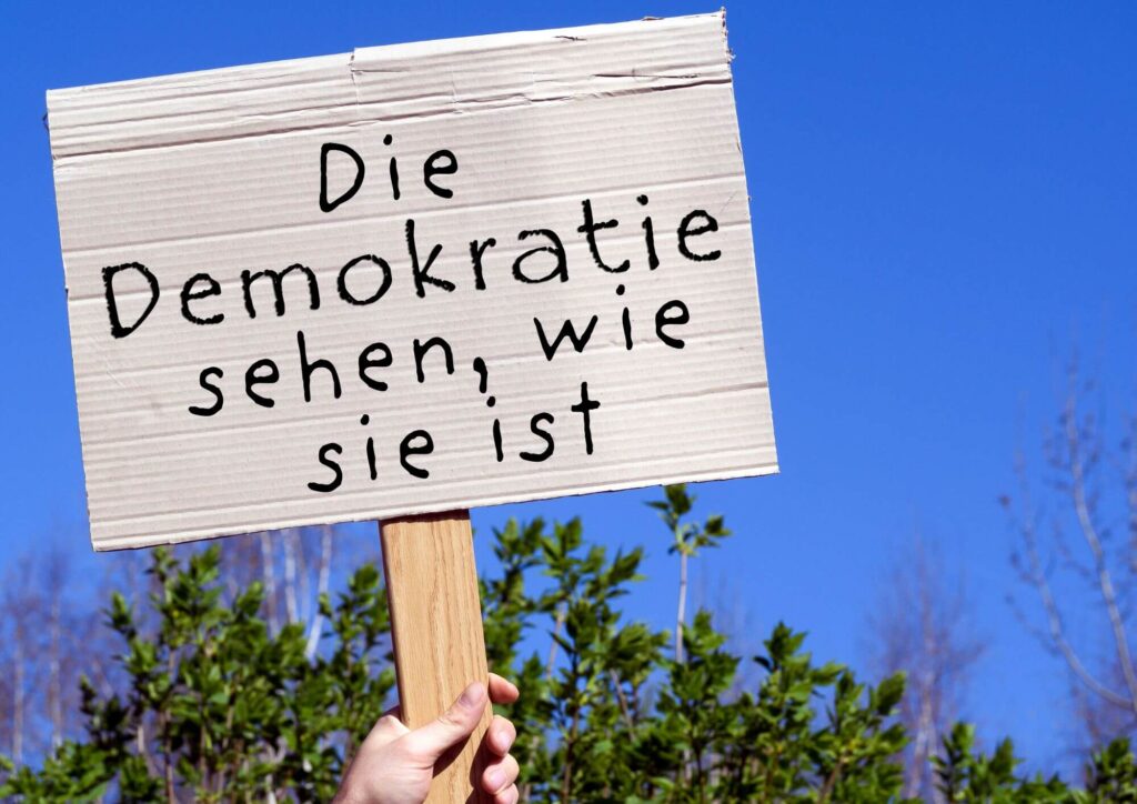 Auf einem hochgehaltenen Pappschild steht: Die Demokratie sehen, wie sie ist